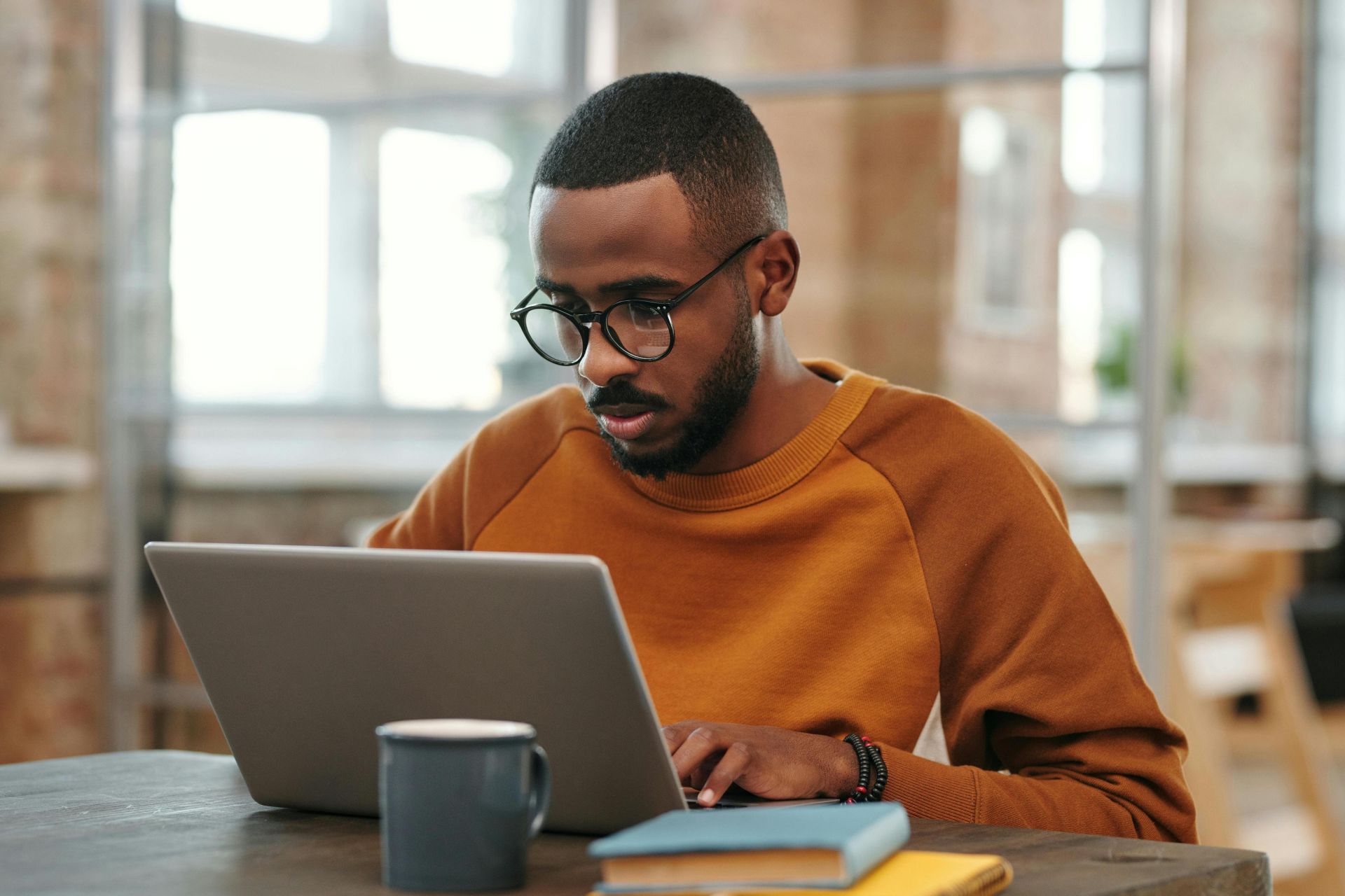 Young man with glasses works on laptop at a wooden table indoors. Warm, productive atmosphere.